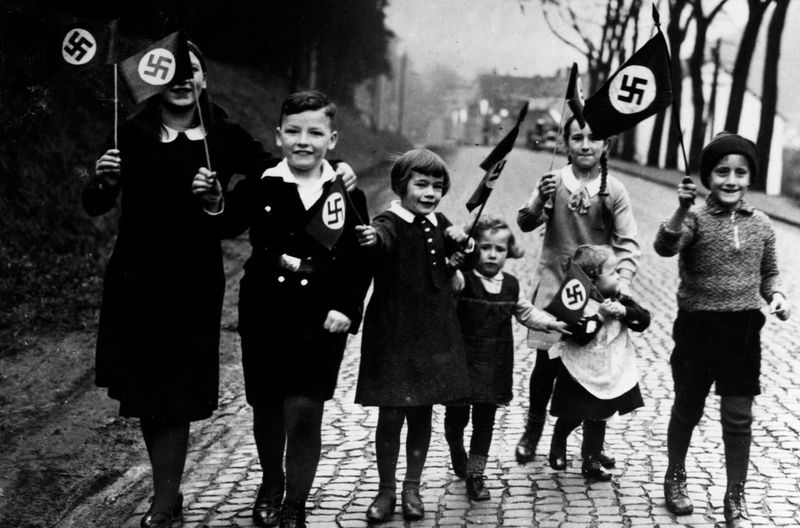 Ein Propagandafoto für die Presse: Kinder gehen mit Hakenkreuzfahnen in der Hand spazieren. 
