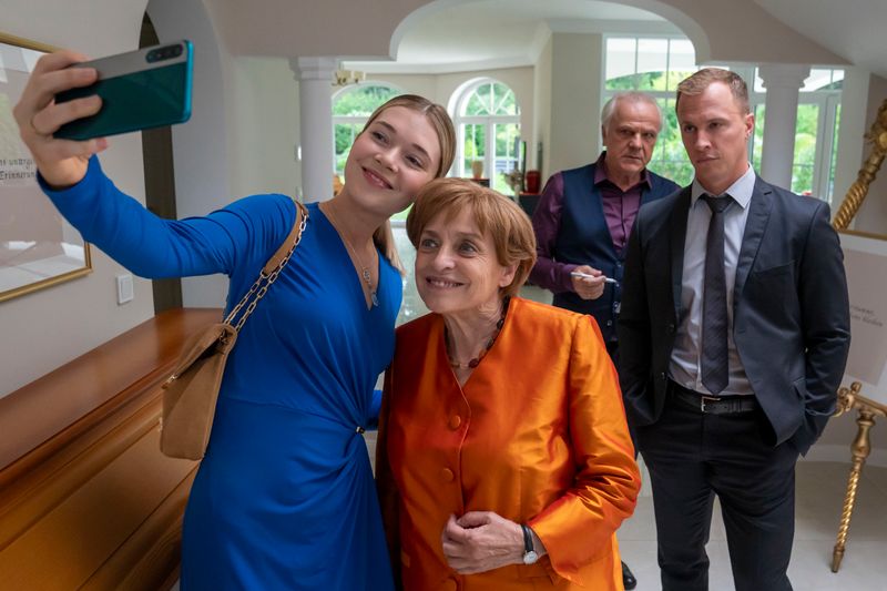 Charu Borscht (Farina Flebbe, links), die junge Frau des zweiten Bestatters im Ort, macht ein Selfie mit Angela Merkel (Katharina Thalbach). Ehemann und Bestatter Borscht (Bernhard Schütz) sowie Merkels Personenschützer Mike (Tim Kalkhof, rechts) schauen zu.
