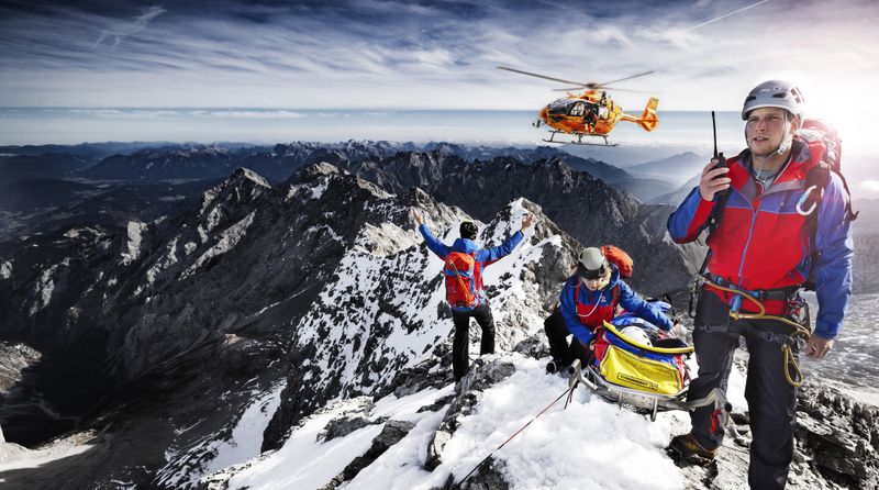 Auch die zweite Staffel der "Bergretter im Einsatz" zeigt Bergwachteinsätze in den bayerischen Alpen - hautnah und in beeindruckenden Bildern. Drei Angehörige der Bergwacht, die sich mit einer geretteten Person auf einem Berggipfel befinden, weisen einen anfliegenden Hubschrauber ein.